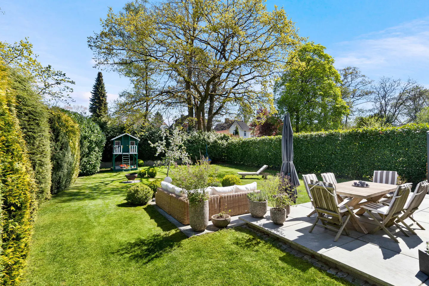A lush green backyard with a patio, wicker furniture, a wooden table with chairs, and a green treehouse.