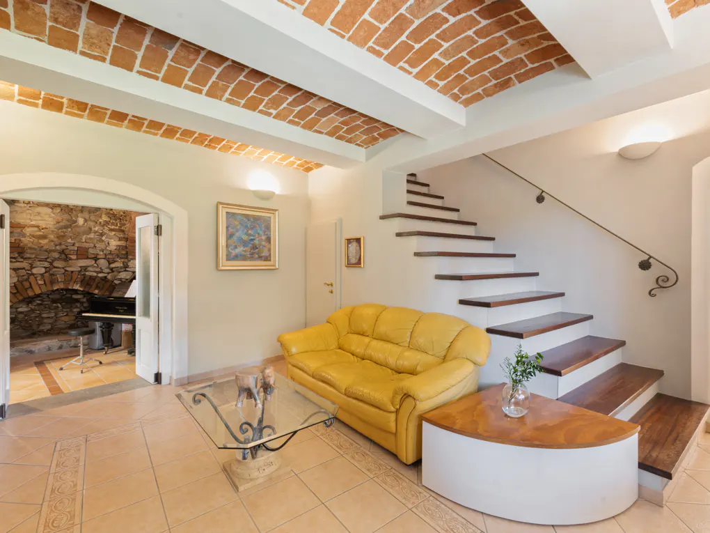 Living room with yellow sofa, glass table, and stairs. Brick ceiling and stone wall visible.