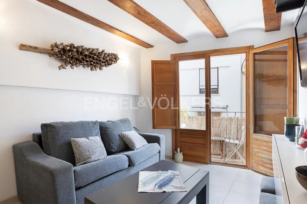 Living room with gray sofa, dark table, and wooden beams. Balcony with open doors. Wall art and Engel & Völkers logo.