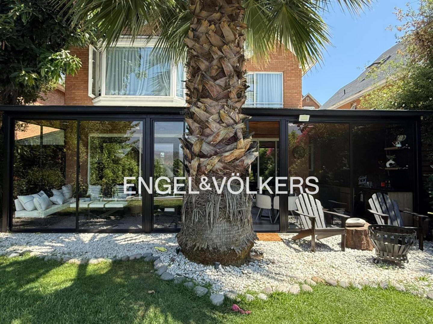 A brick house with a glass-enclosed patio, a large palm tree in the foreground, and outdoor seating on a gravel yard.