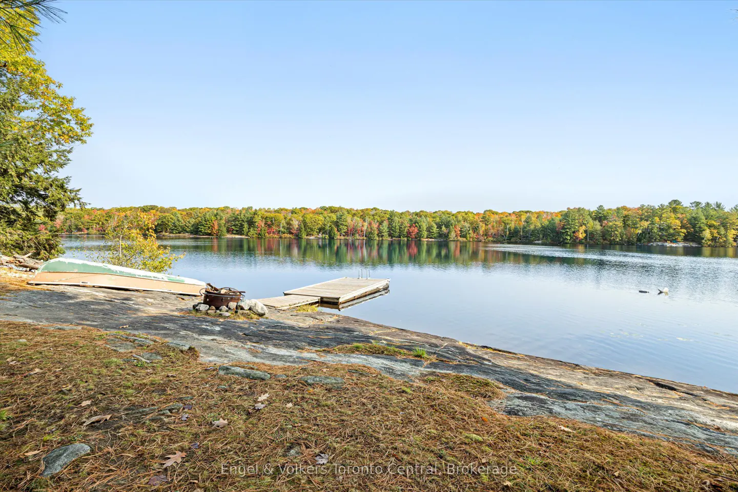 Lake view with a dock, fire pit, and overturned boat on a rocky shore. Trees with fall foliage line the opposite shore under a clear blue sky.