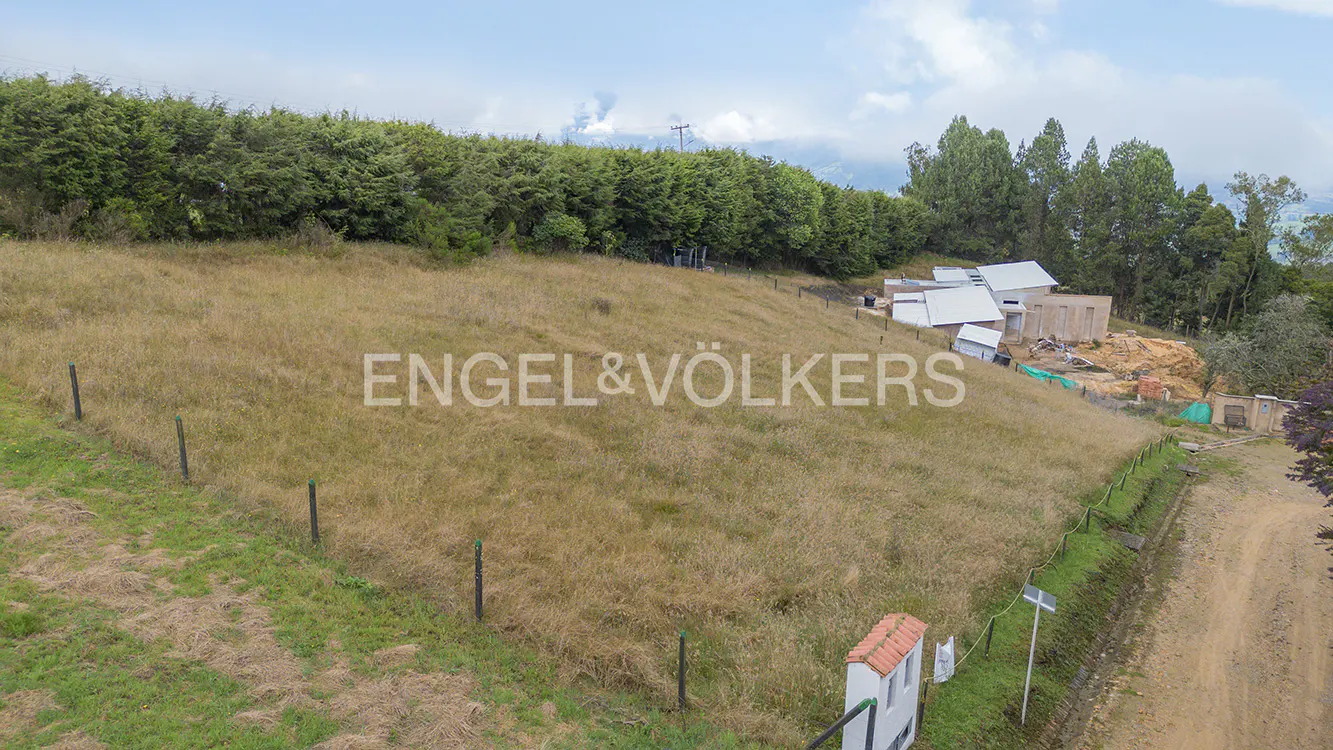 A hillside lot with tall, dry grass, a fence, and a partially constructed house in the background.
