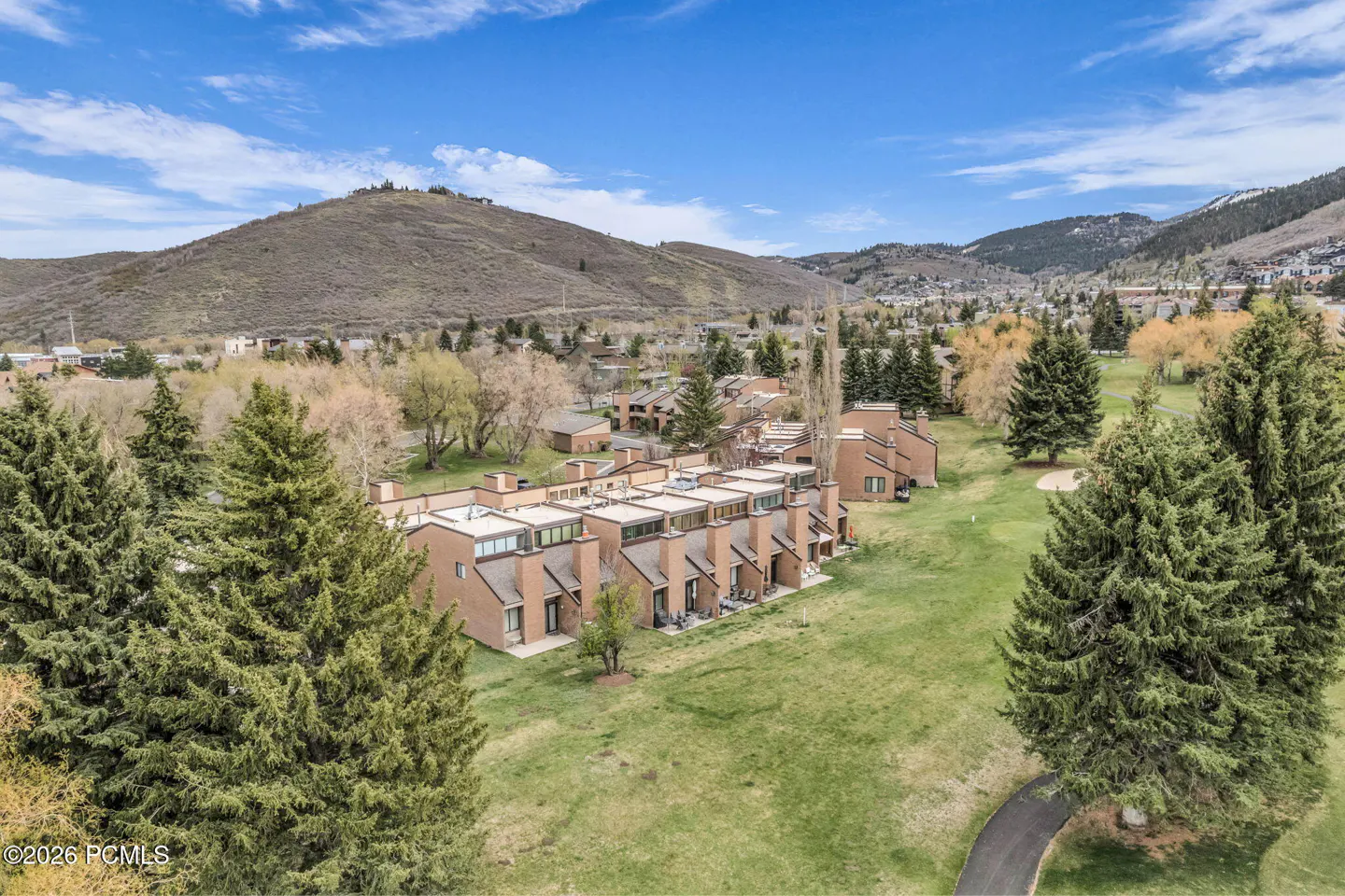 Aerial view of brown townhouses on a green lawn, with mountains and blue sky in the background.