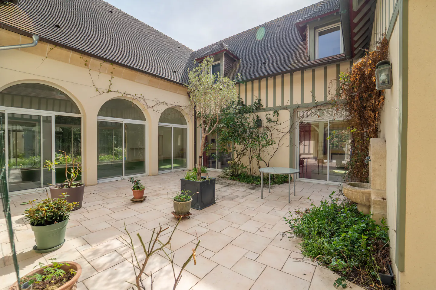 Courtyard view with potted plants, stone tiles, and arched windows. A small table sits near a wall with climbing vines and a stone fountain.