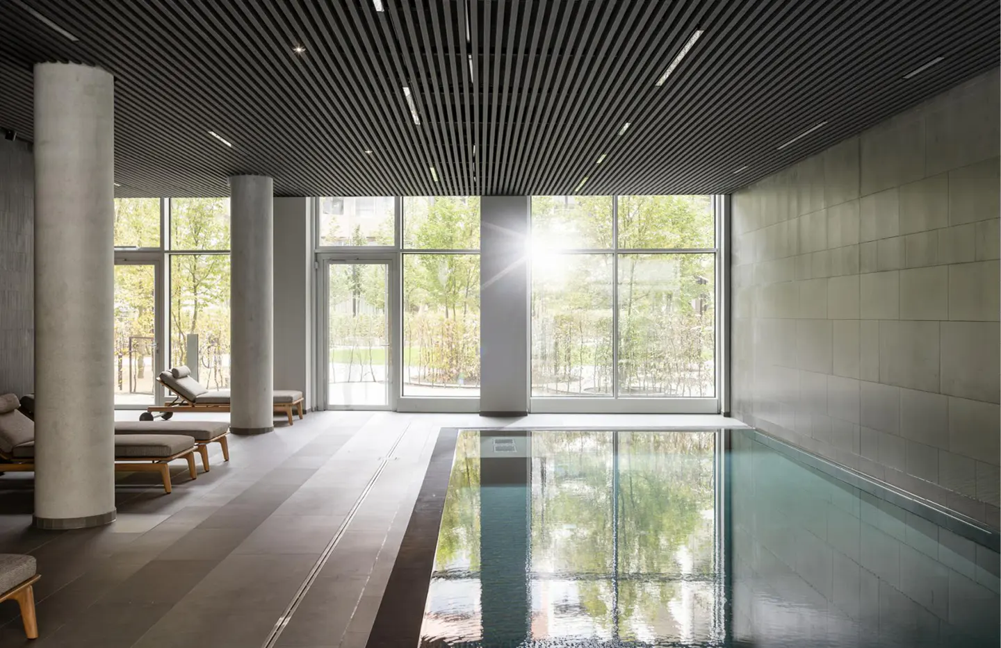 Indoor swimming pool with lounge chairs, concrete pillars, and floor-to-ceiling windows overlooking a sunny garden.