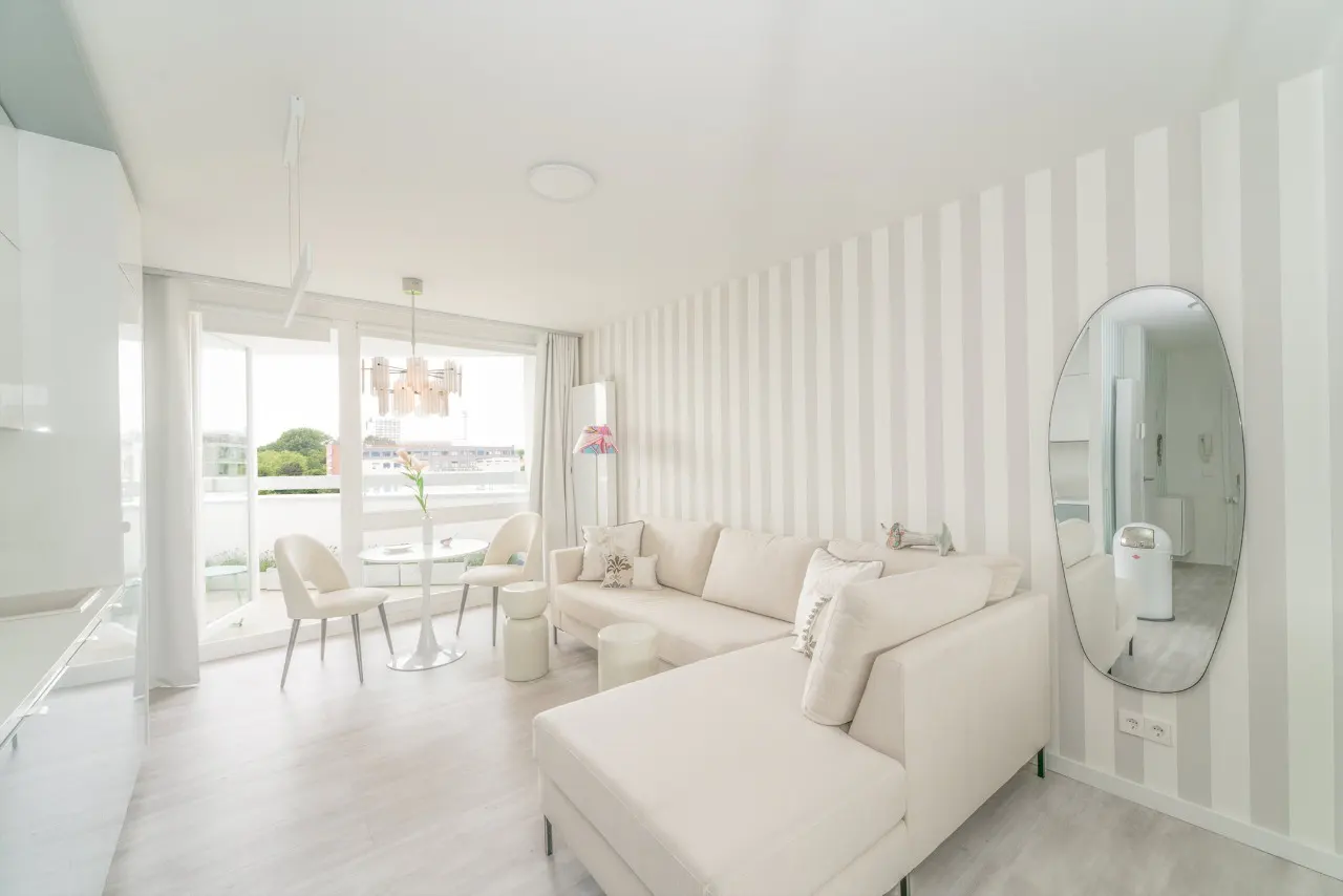 Bright living room with a white sectional sofa, striped wallpaper, a dining area by the balcony, and an oval wall mirror.