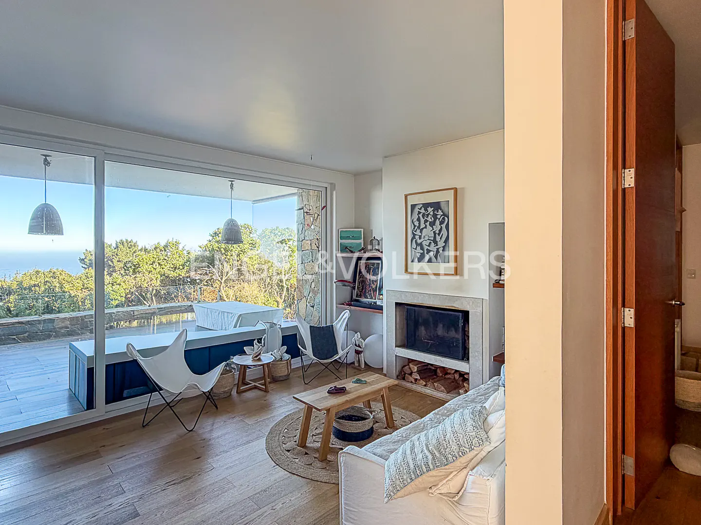 Bright living room with hardwood floors, white sofa, fireplace, and large windows overlooking trees and the ocean.