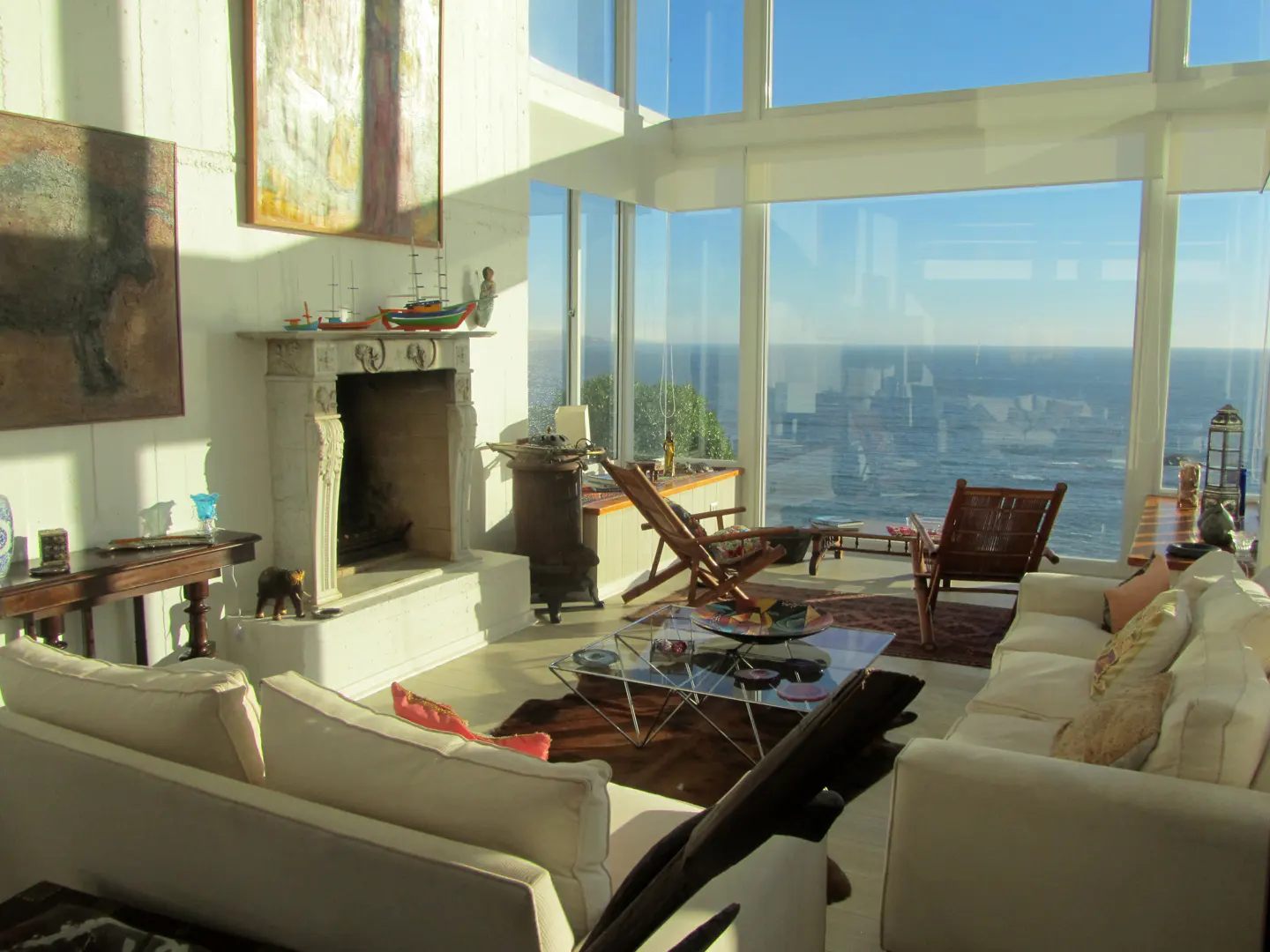 Bright living room with white sofas, fireplace, and floor-to-ceiling windows overlooking the ocean. Art adorns the walls.