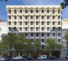Exterior of a multi-story beige building with dark windows and retail shops on the ground floor. Trees line the street.