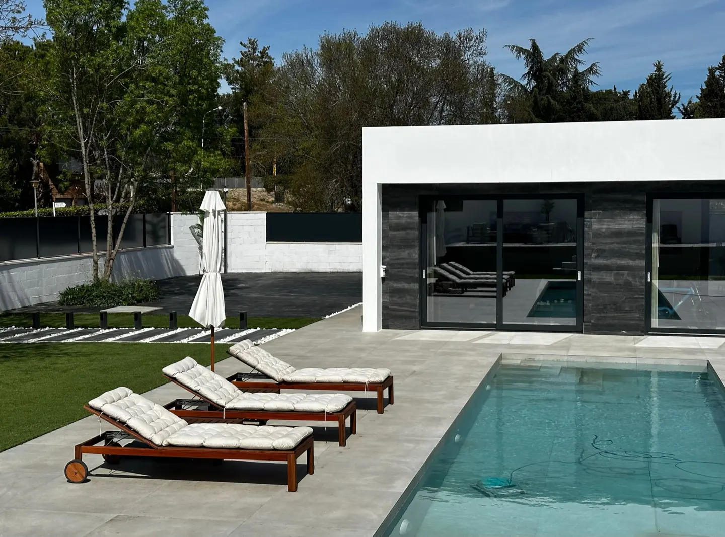 Modern home exterior with a pool, lounge chairs, and a white umbrella. Trees and a blue sky are in the background.