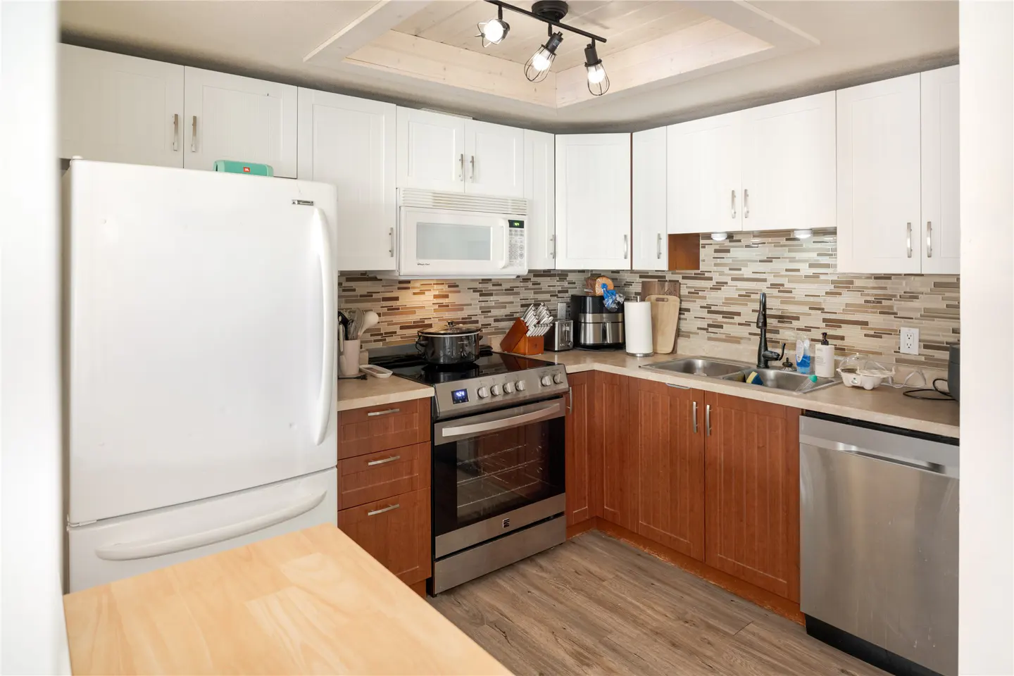 A kitchen with white upper cabinets, wood lower cabinets, stainless steel appliances, and a mosaic tile backsplash.