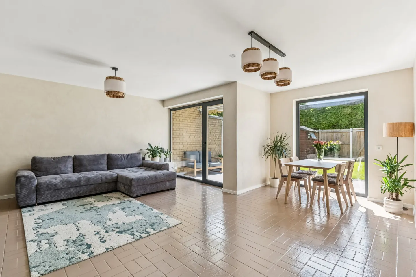 A bright, open-plan living and dining area with a gray sectional sofa, a dining table with four chairs, and large windows overlooking a garden.