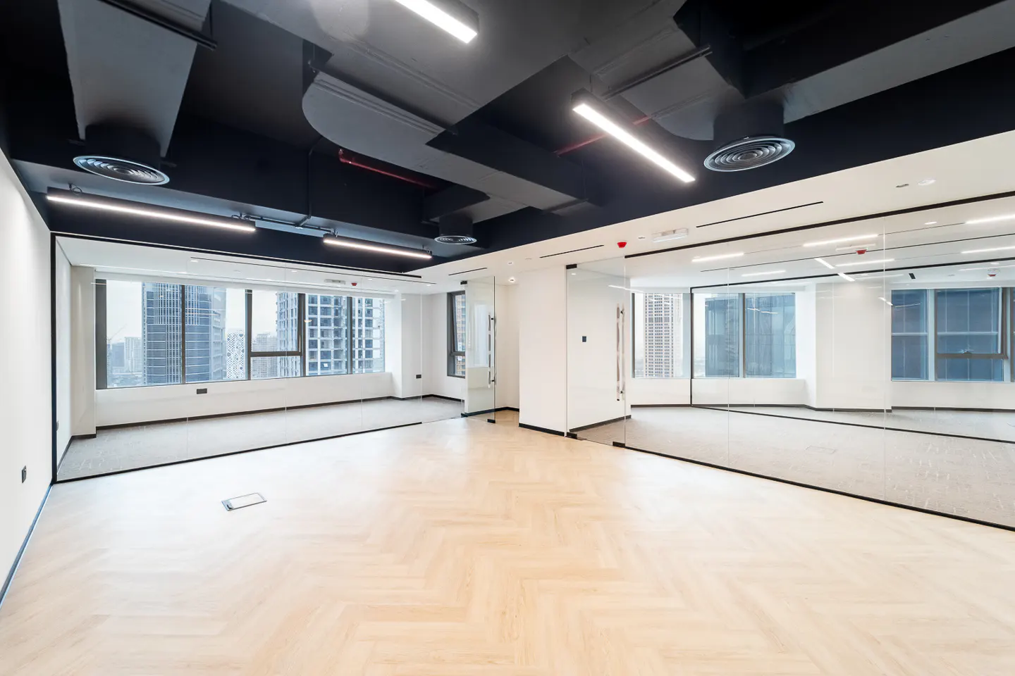 An empty office space with herringbone wood floors, white walls, and large windows overlooking a city skyline. The ceiling is black with modern lighting.