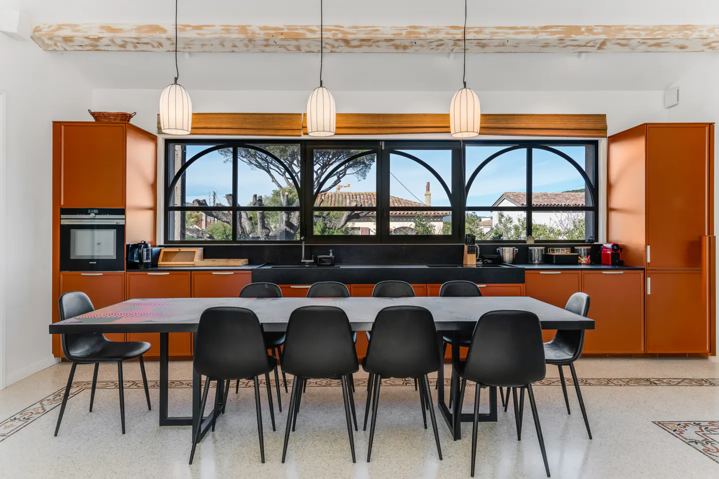 A modern kitchen with orange cabinets, a black countertop, and a long black table with eight chairs. Large arched windows offer a view of the outdoors.