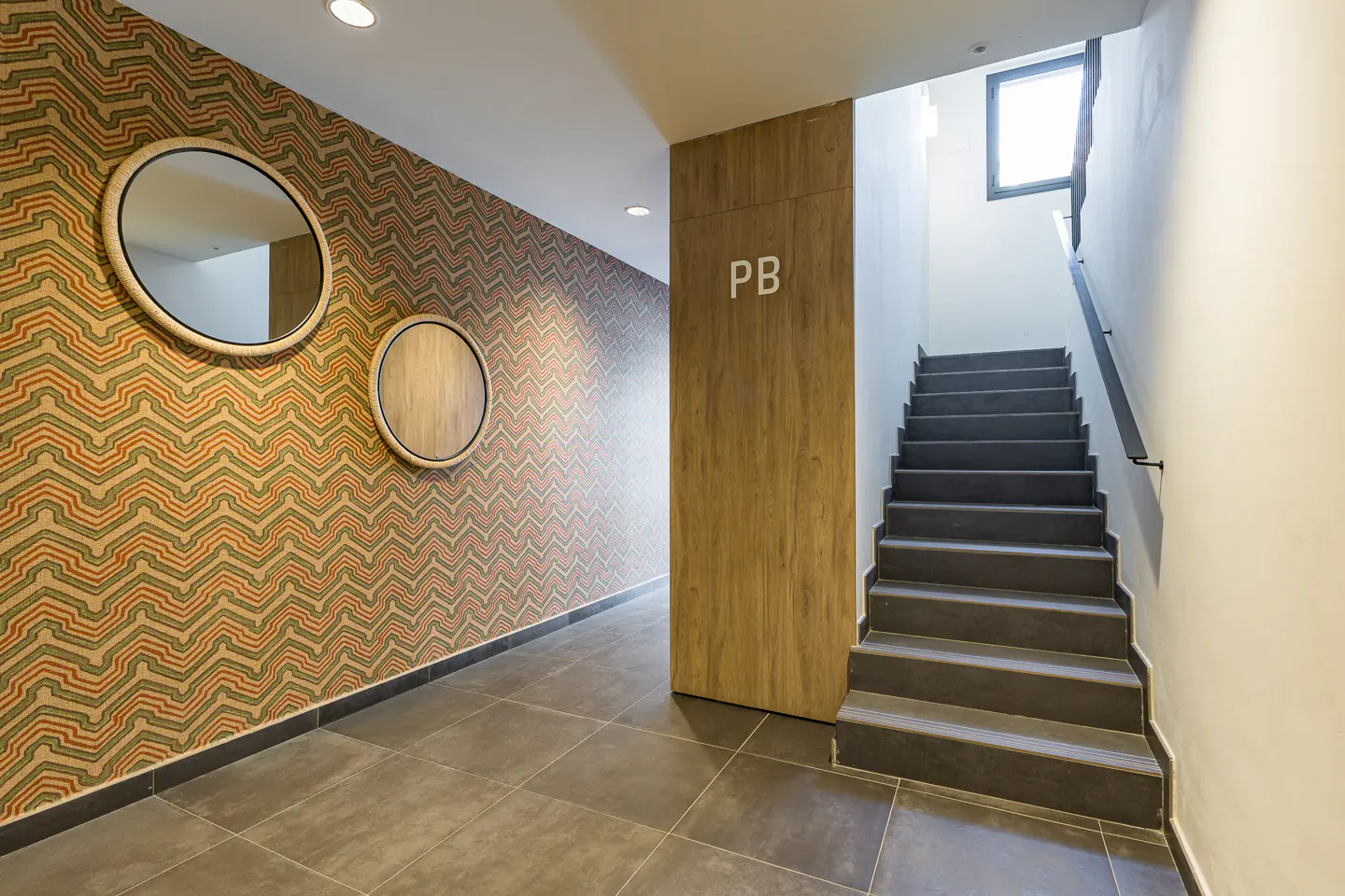 Hallway with patterned wallpaper, round mirrors, and gray tile floor. A wooden panel with "PB" leads to a staircase with a window.
