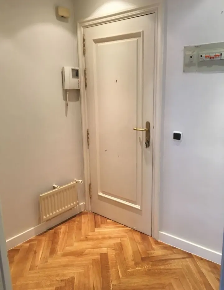 Entryway with herringbone wood floors, white walls, and a white door with brass hardware. A radiator and intercom are on the left wall.