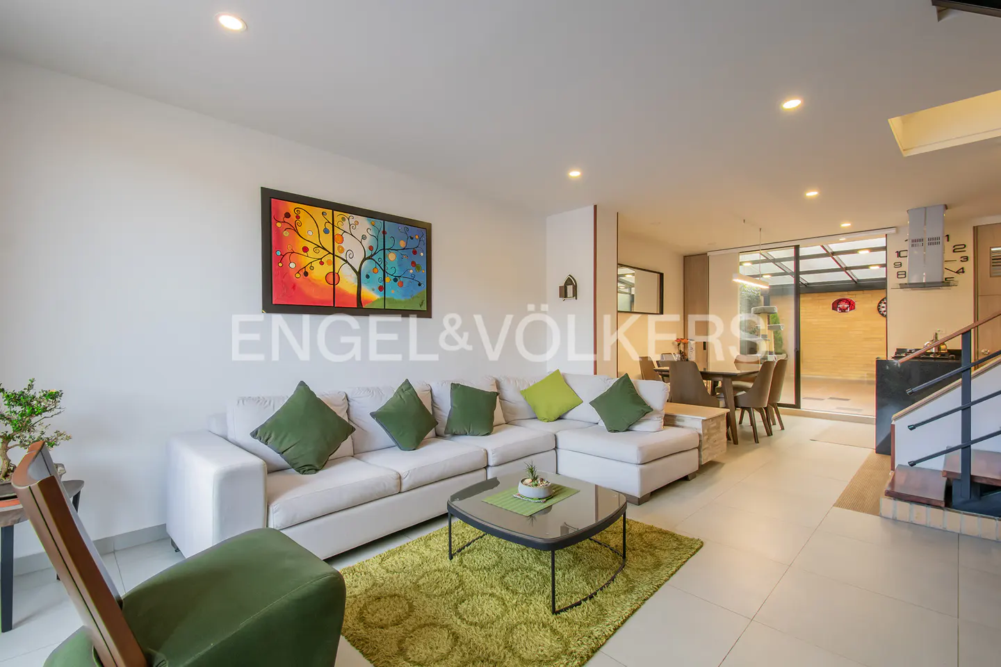 Bright living room with white sectional sofa, green pillows, and a colorful tree painting. A green rug sits under a glass coffee table.