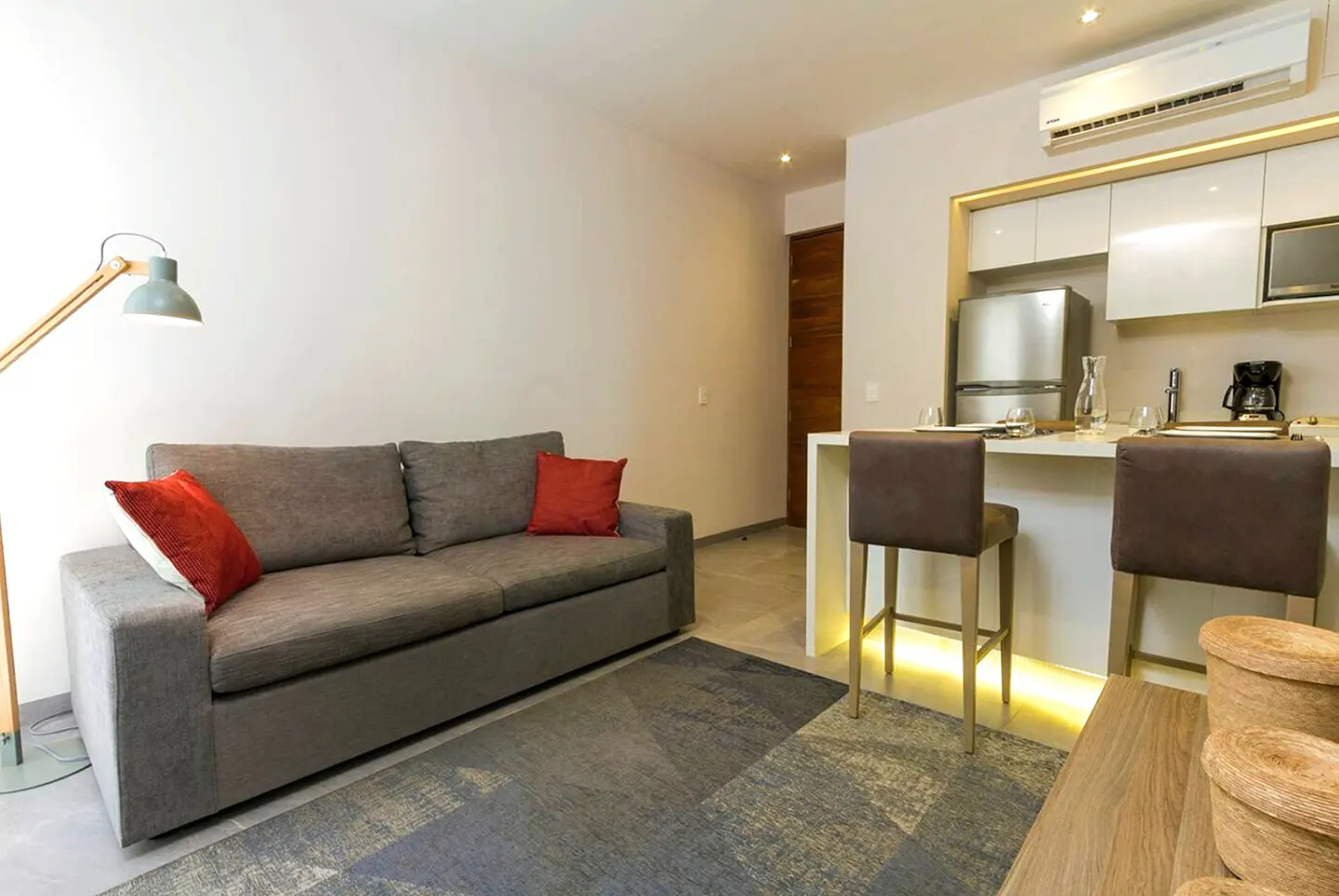 Living space with gray sofa, red pillows, floor lamp, and kitchen with bar stools.
