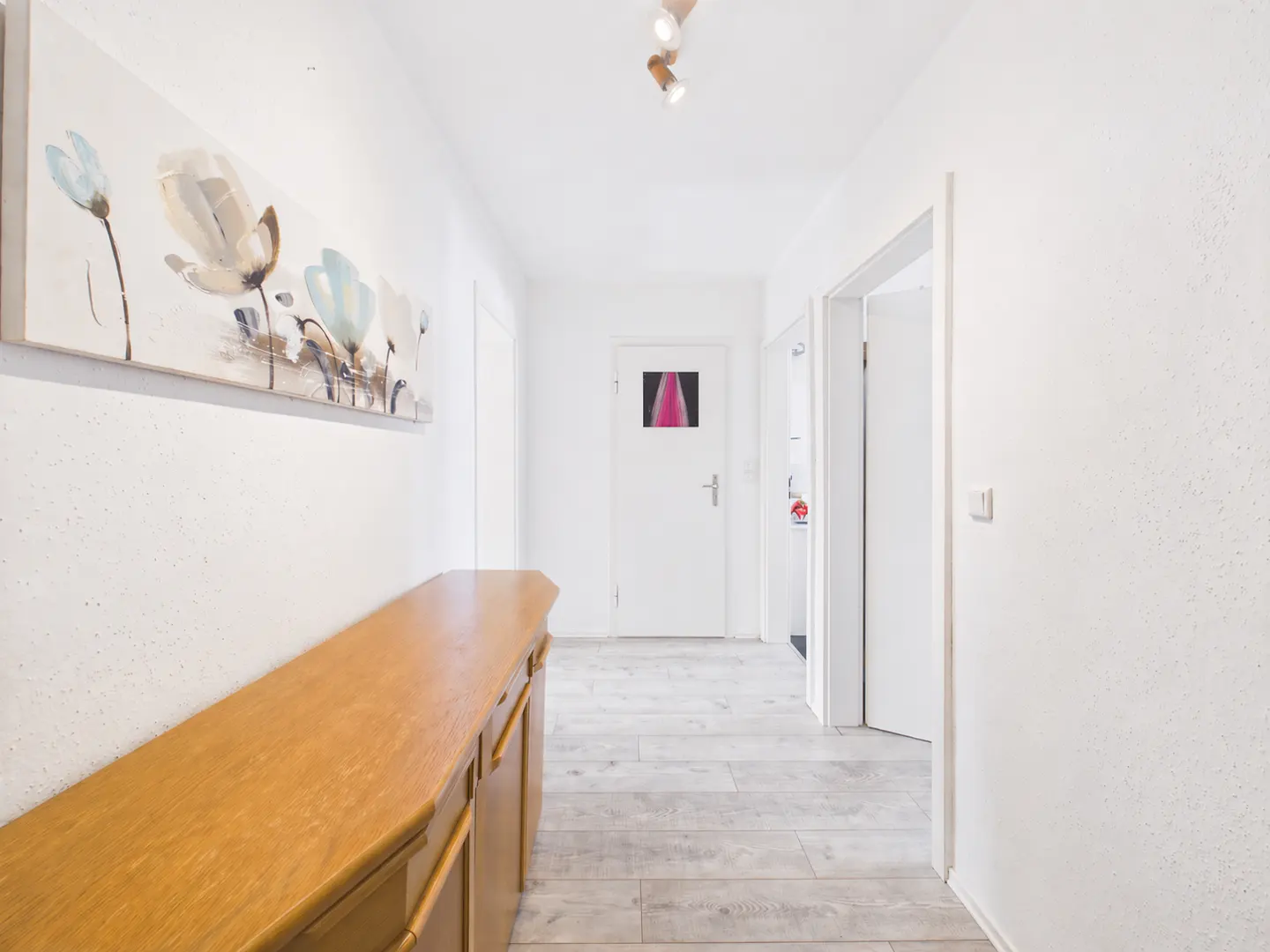 Hallway with white walls, light wood floors, and a long wooden cabinet. A floral painting hangs above the cabinet. A white door is at the end of the hall.