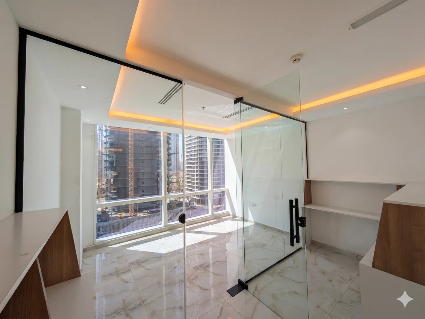 Bright office space with glass-walled room, marble floors, and city view. White walls, built-in desk and shelves.