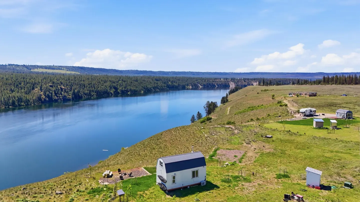 Aerial view of a white tiny house with a gray roof, overlooking a blue river and green landscape.