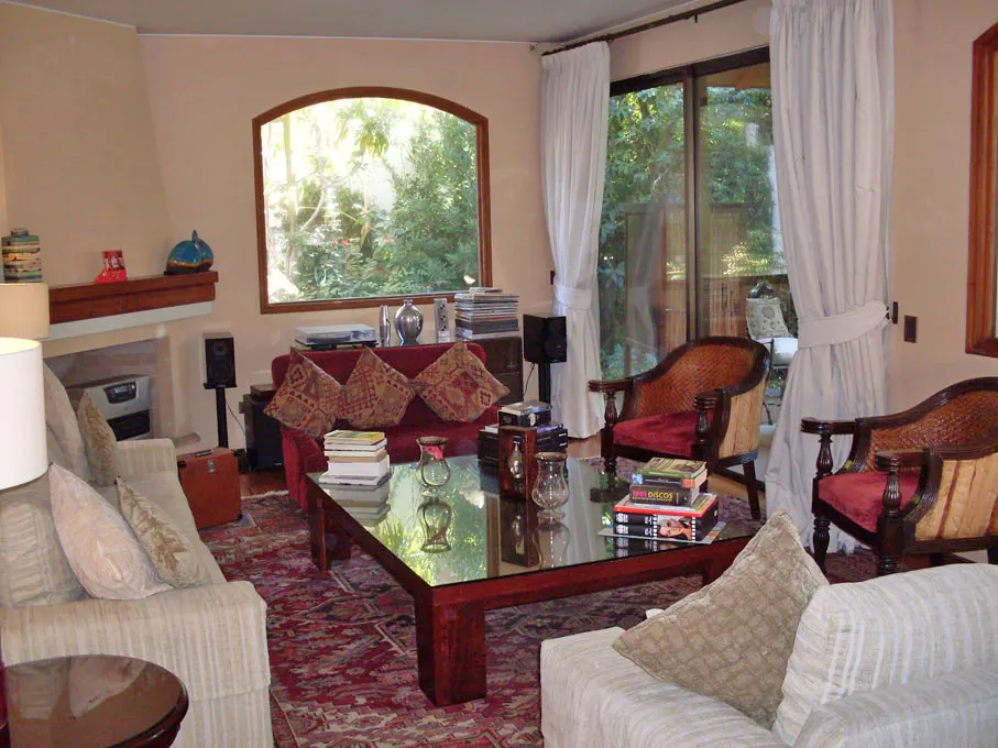 Living room with beige walls, a red patterned rug, and a glass coffee table. There are two sofas, two chairs, and a fireplace. A large window looks out onto a green garden.