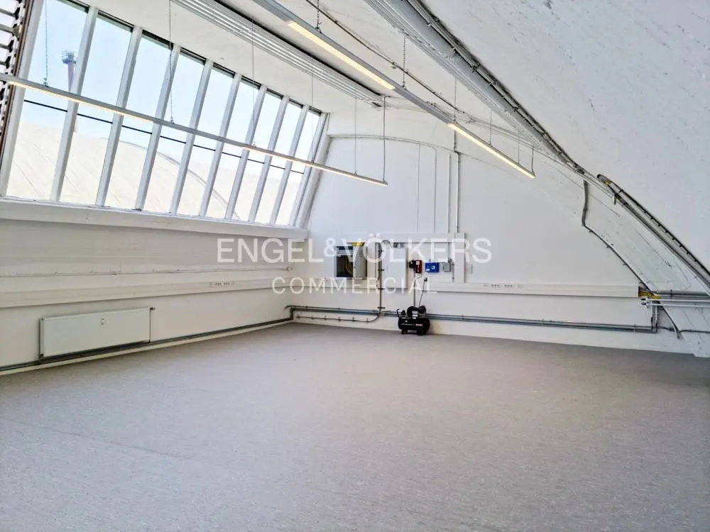 Bright, empty commercial space with white walls, gray floor, and large windows. Fluorescent lights hang from the ceiling. Engel & Volkers logo visible.