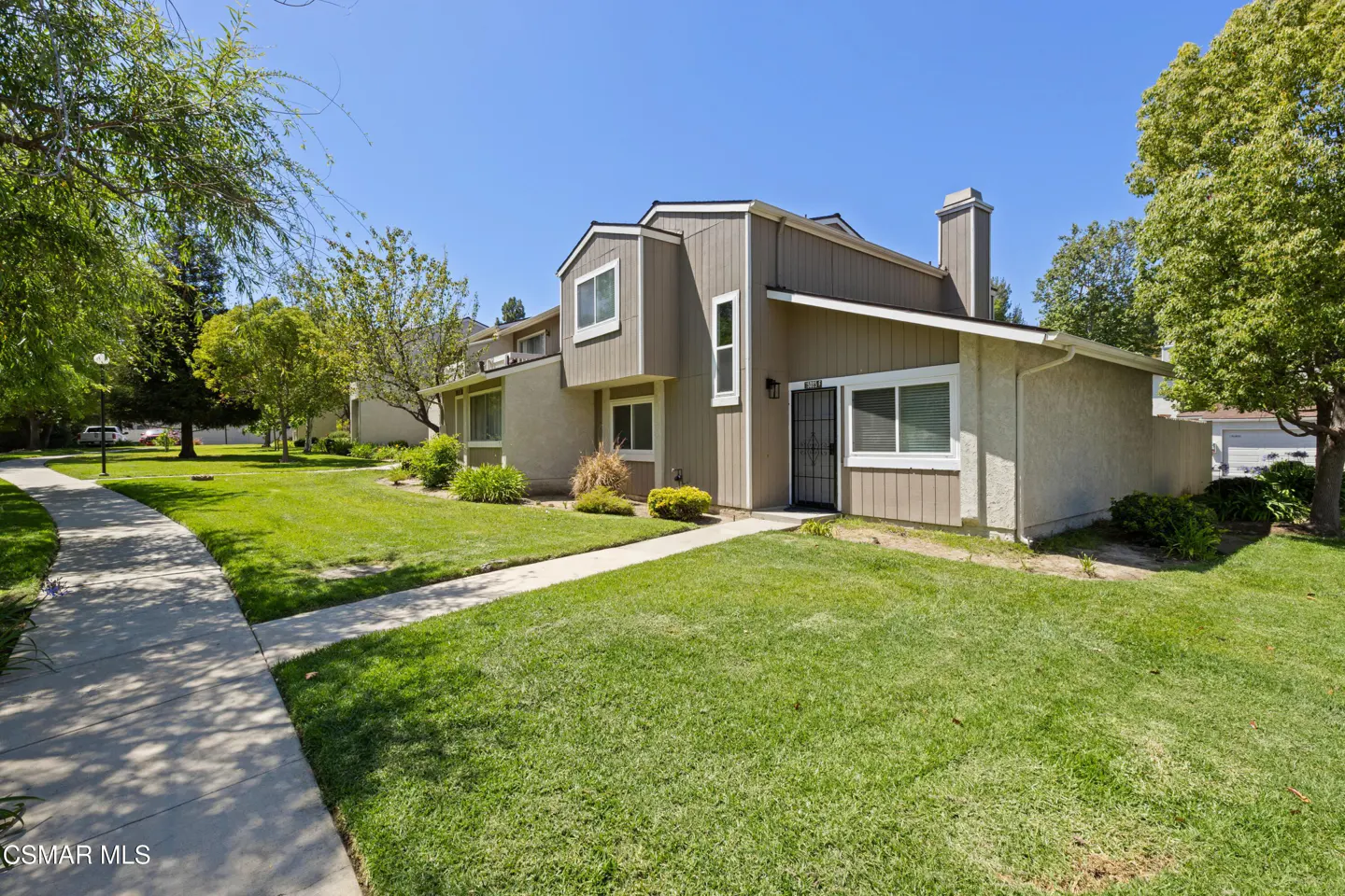 Exterior view of a two-story townhouse with beige siding, green lawn, and a concrete walkway on a sunny day.