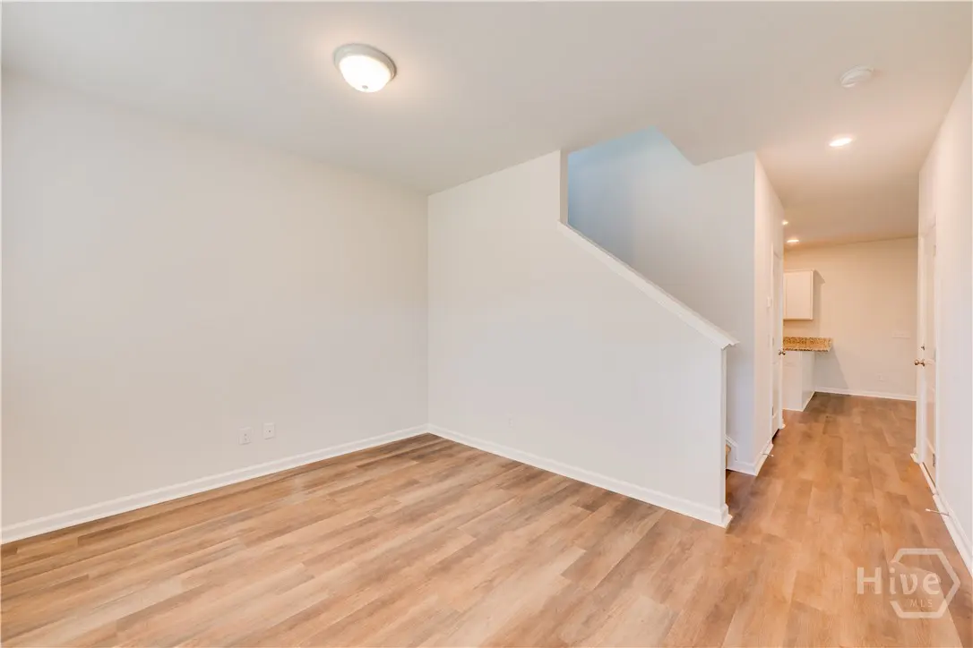 Bright, empty room with wood floors and white walls. Stairs are visible. Hallway leads to a small kitchen area.