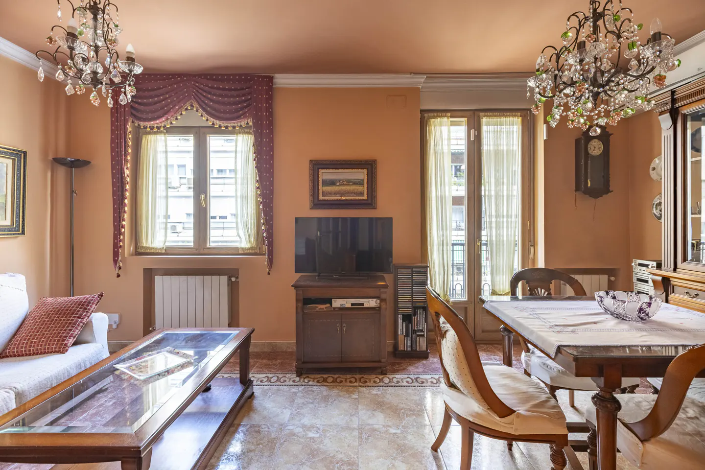 A living and dining room with peach walls, chandeliers, and a glass-topped coffee table.