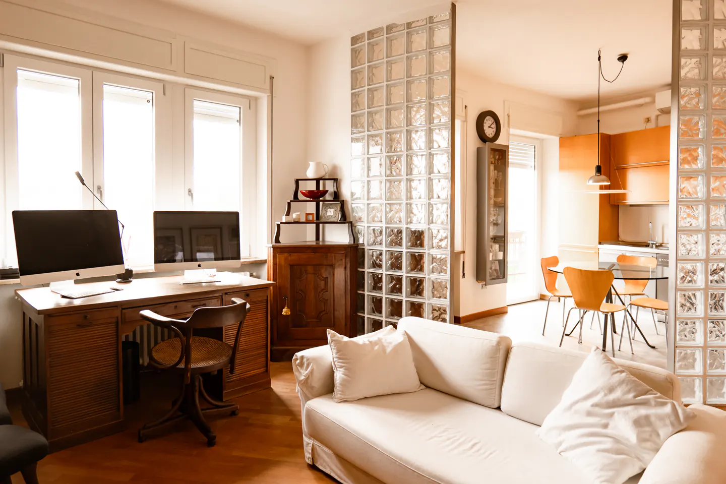 Bright living room with a white sofa, wooden desk with two computers, and a glass block wall separating the kitchen.