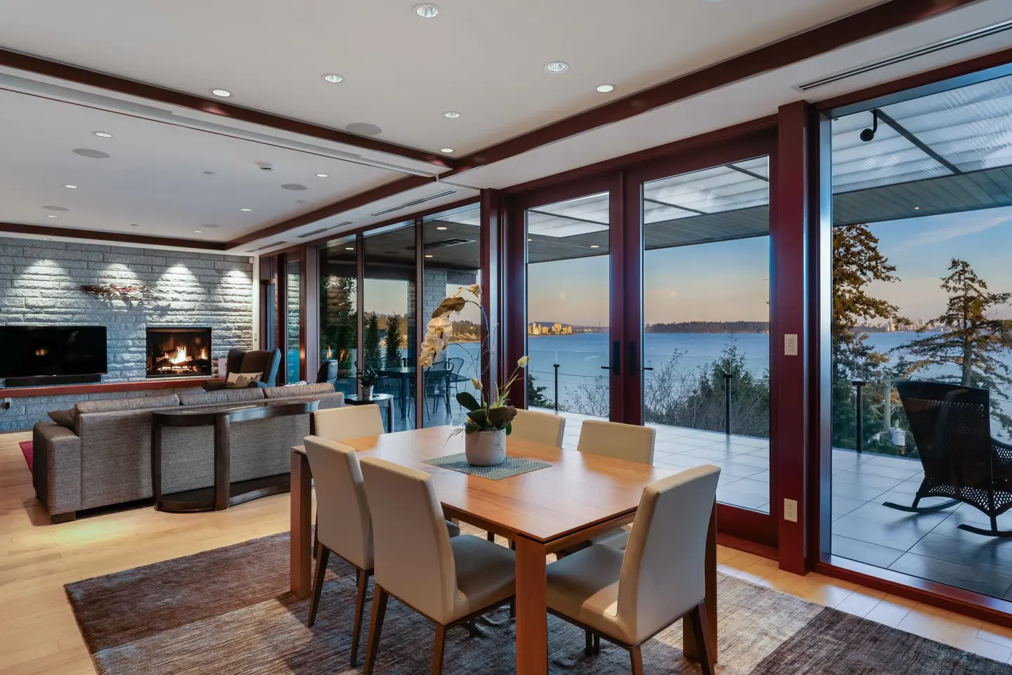 Open-concept living and dining area with a stone fireplace, wood table, and sliding glass doors to a patio with a water view.