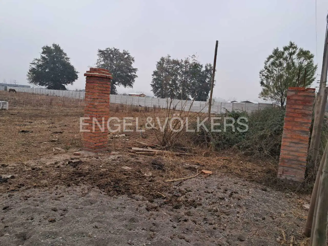 Vacant lot with brick pillars and Engel & Völkers logo. A gray sky and trees are in the background.