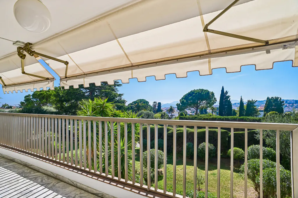 Balcony view with a beige awning, metal railing, and a garden with green trees and bushes. Blue sky in the background.