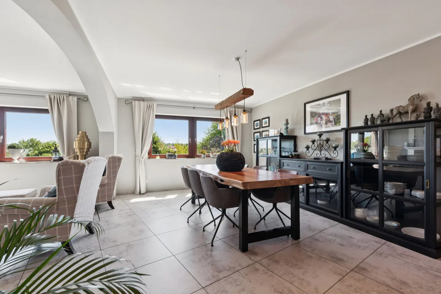 Bright dining room with a wood table, gray chairs, and a black cabinet. Windows with white curtains let in natural light.