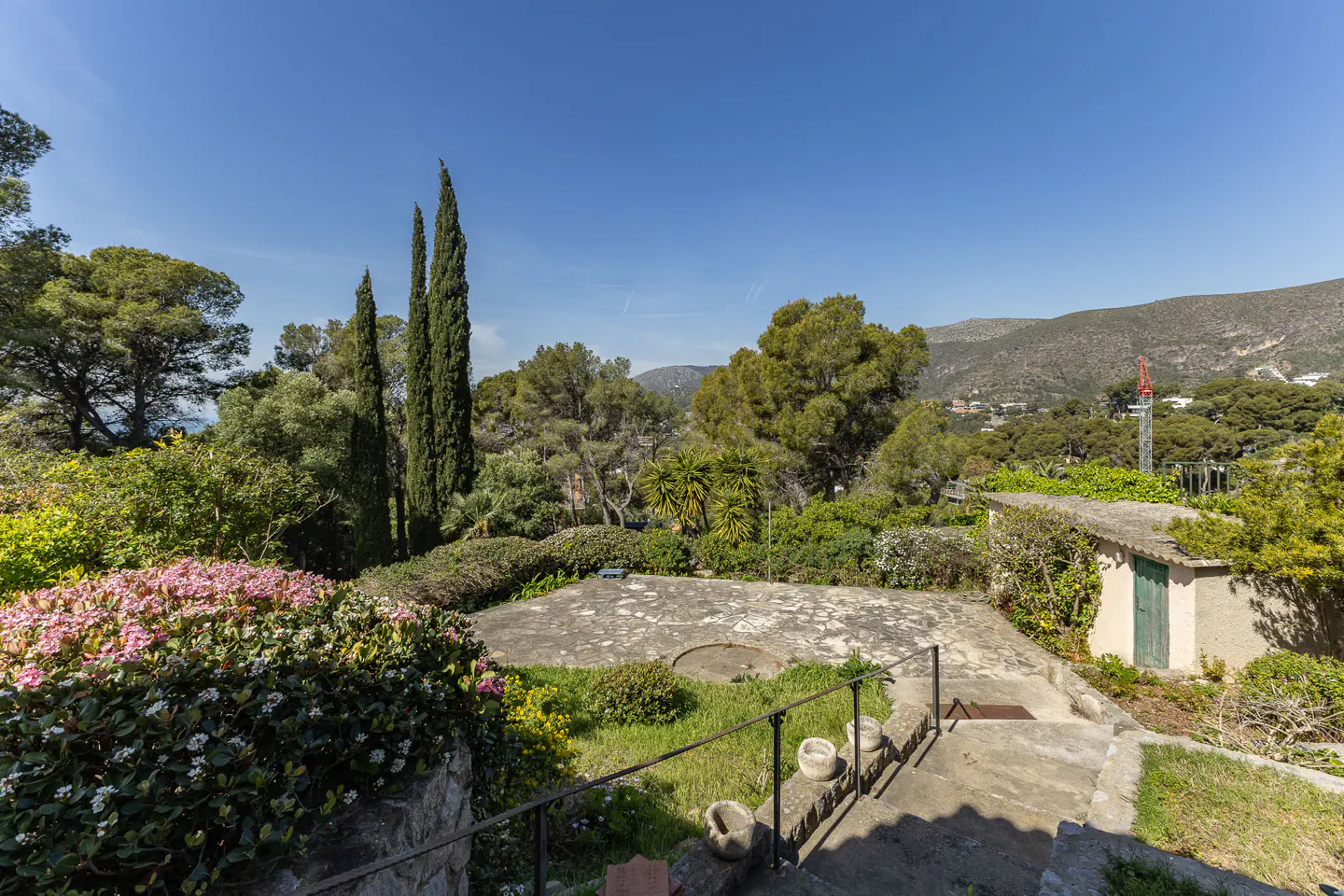 A garden view with stone steps, a metal railing, and lush greenery under a clear blue sky. A small shed is visible.