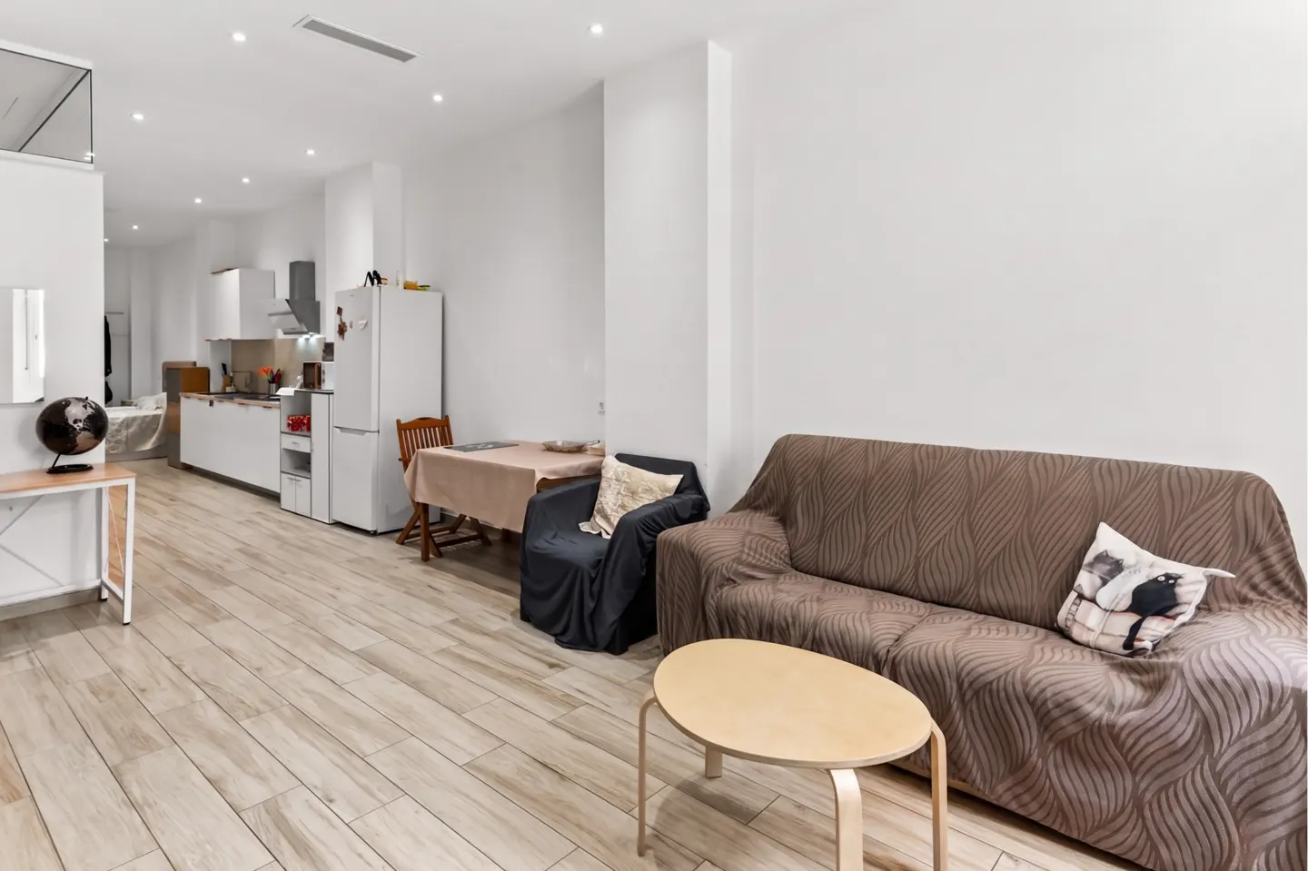 Bright, open-concept apartment with wood floors, white walls, and recessed lighting. Features a brown sofa, kitchen, and dining area.
