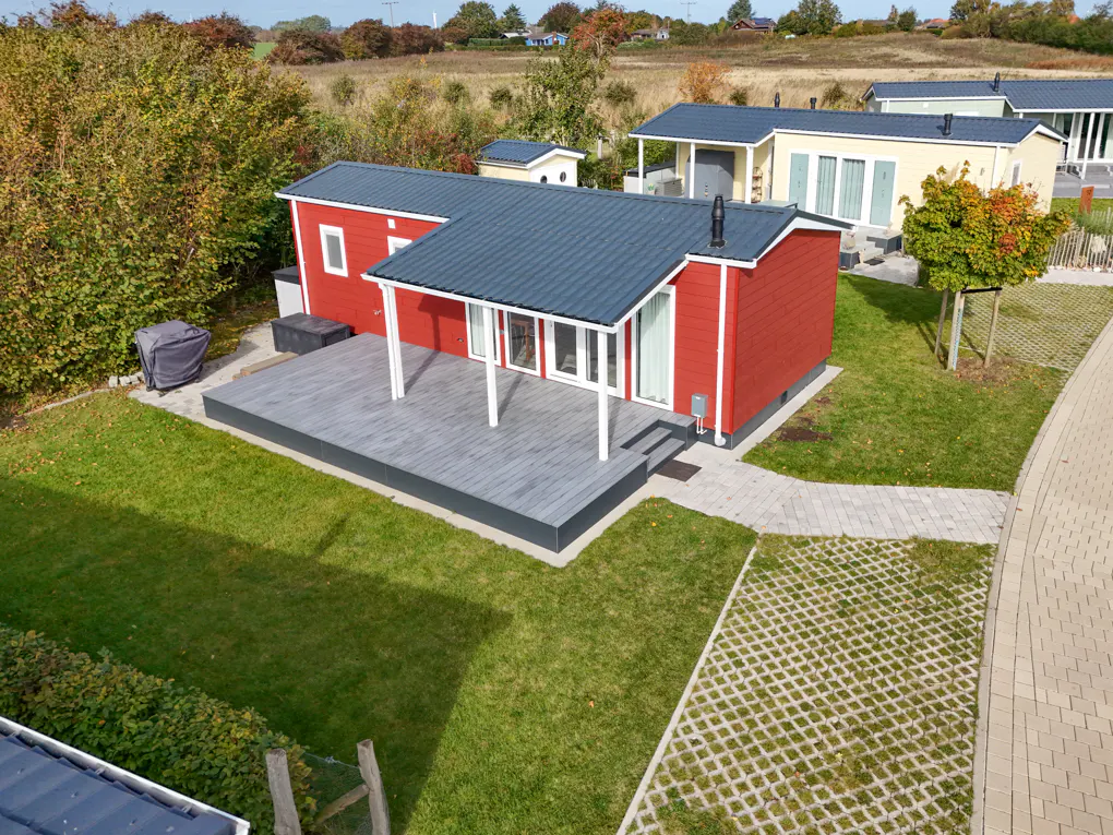 Aerial view of a red house with a gray roof and deck, surrounded by green grass and other houses.