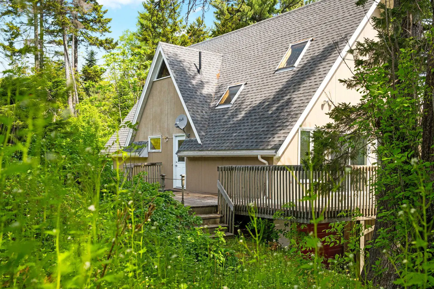 A tan A-frame house with a gray shingled roof and wooden deck, nestled in a lush, green wooded setting.