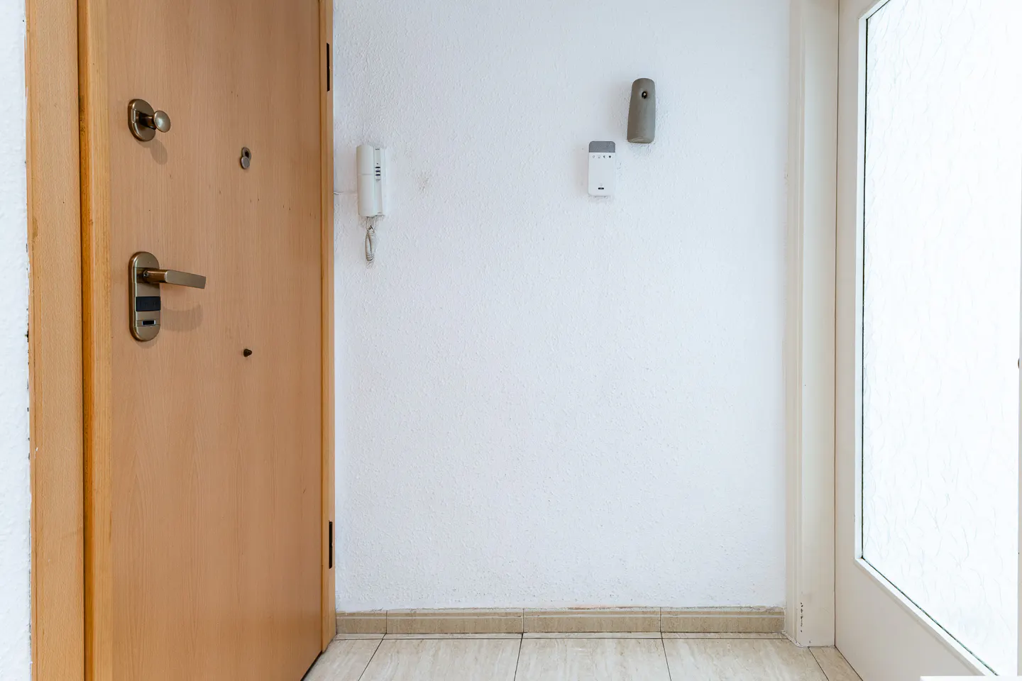 Entryway with a light wood door, white walls, and beige tile floor. Intercom, thermostat, and air freshener are mounted on the wall.