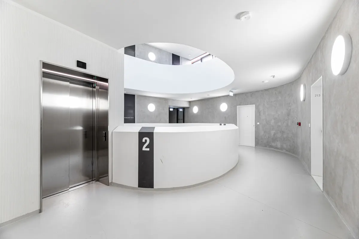 Modern building interior with a stainless steel elevator, white curved wall with the number 2, and a gray textured wall with round lights.