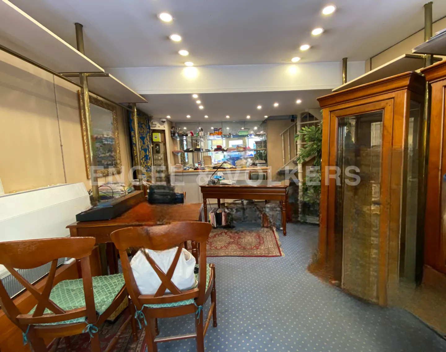 Interior view of a shop with wooden tables, chairs, display cases, and blue carpet. Recessed lighting illuminates the space.