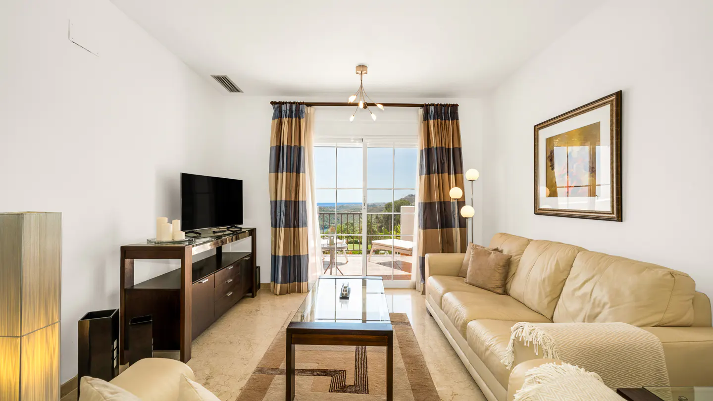 Bright living room with beige sofa, TV, and glass table. Curtains frame a balcony view of green hills. Artwork hangs on the white wall.