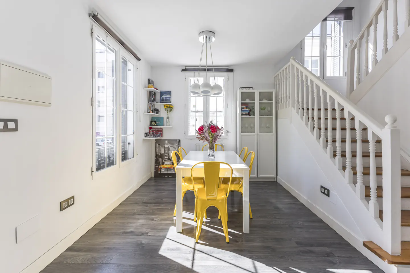 Bright dining room with white walls, gray wood floors, and a white table surrounded by six yellow chairs. A white staircase is on the right.