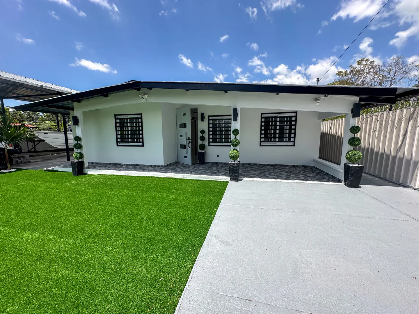 A white, one-story house with black trim, windows, and potted topiary trees. A green lawn and gray driveway lead to the home under a blue sky.