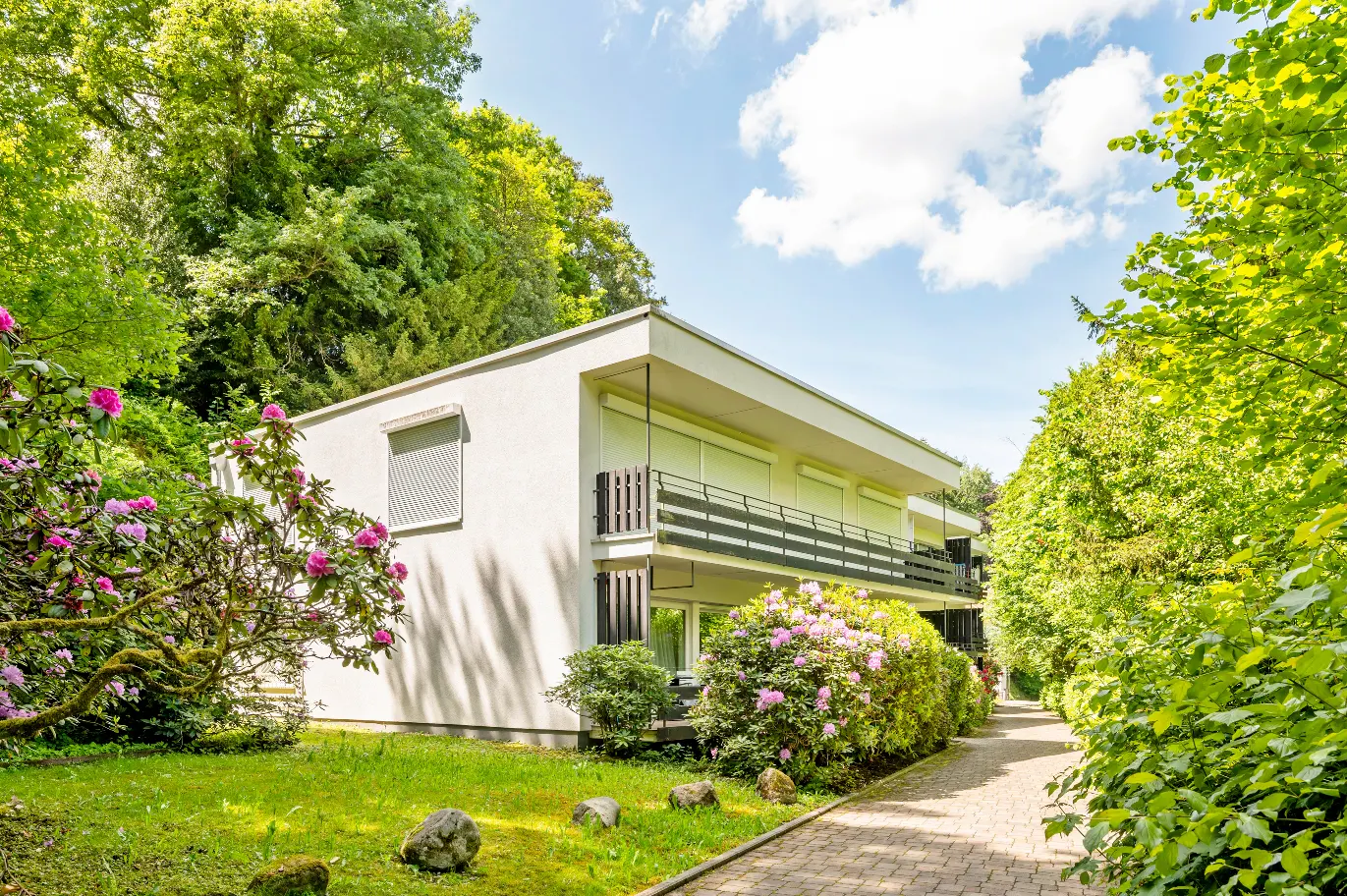 Two-story white apartment building with black balconies, surrounded by green trees and pink rhododendrons. Paved walkway.