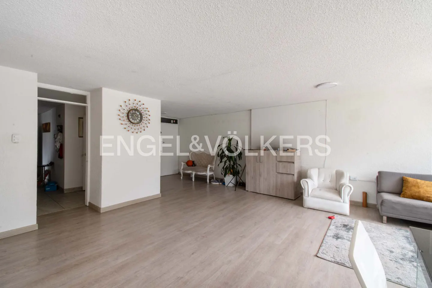 A bright, open living room with light wood floors, white walls, and modern furniture. Engel & Völkers logo is visible.