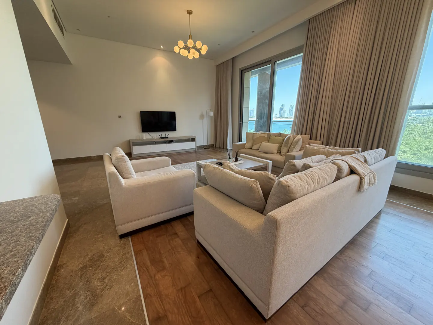 Modern living room with beige sofas, a TV on a console, a gold chandelier, and floor-to-ceiling windows with a water view.