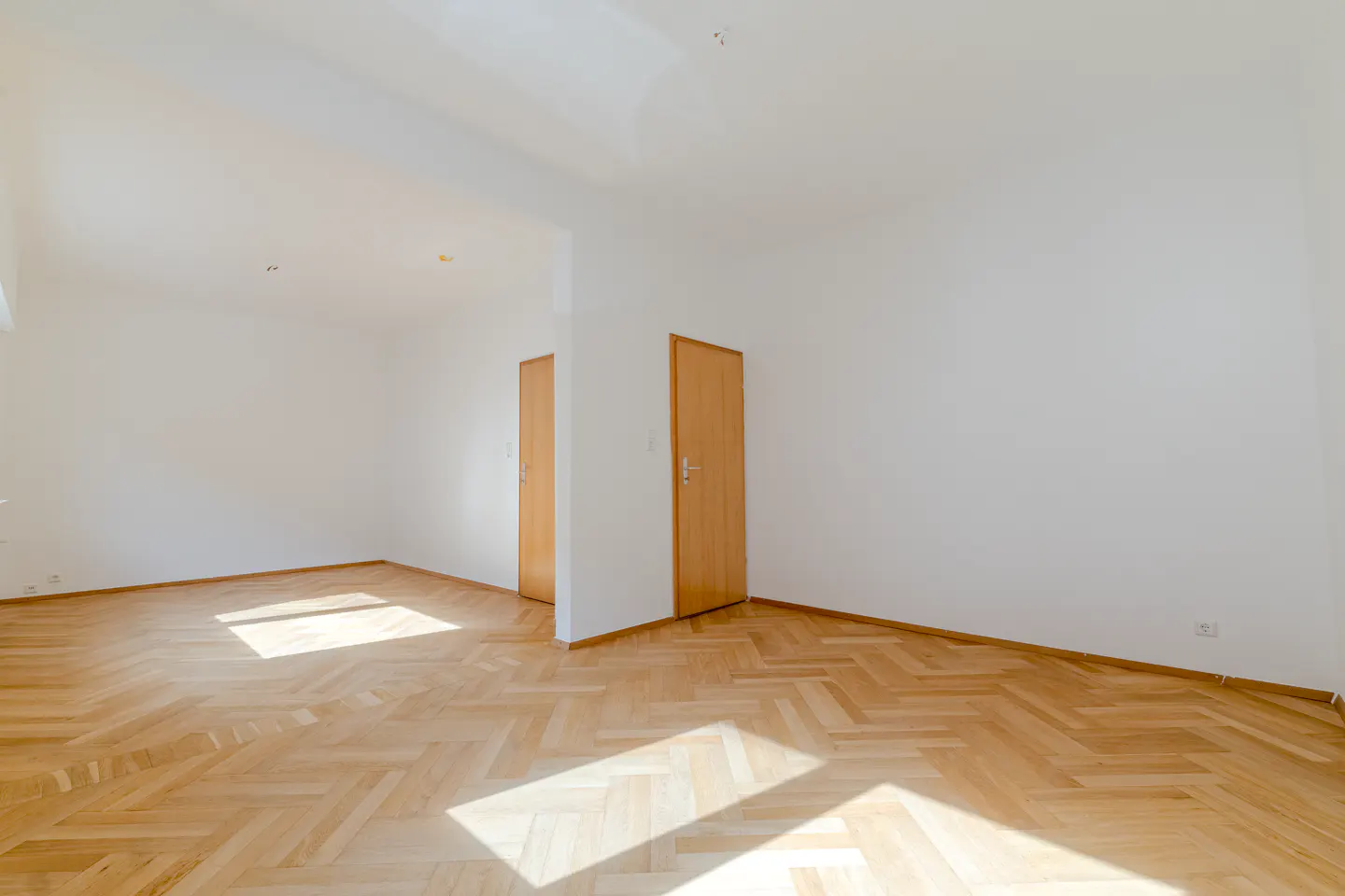 Bright, empty room with herringbone wood floors, white walls, and two light-wood doors. Sunlight streams in from a window.