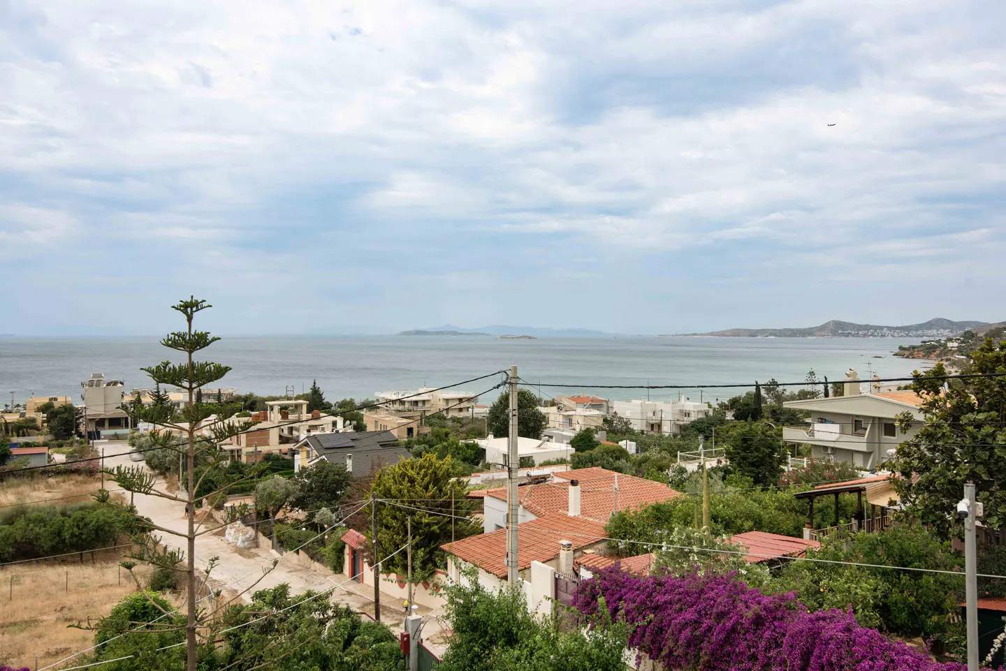 Elevated view of a coastal town with terracotta-roofed houses, lush greenery, purple bougainvillea, and the calm sea.